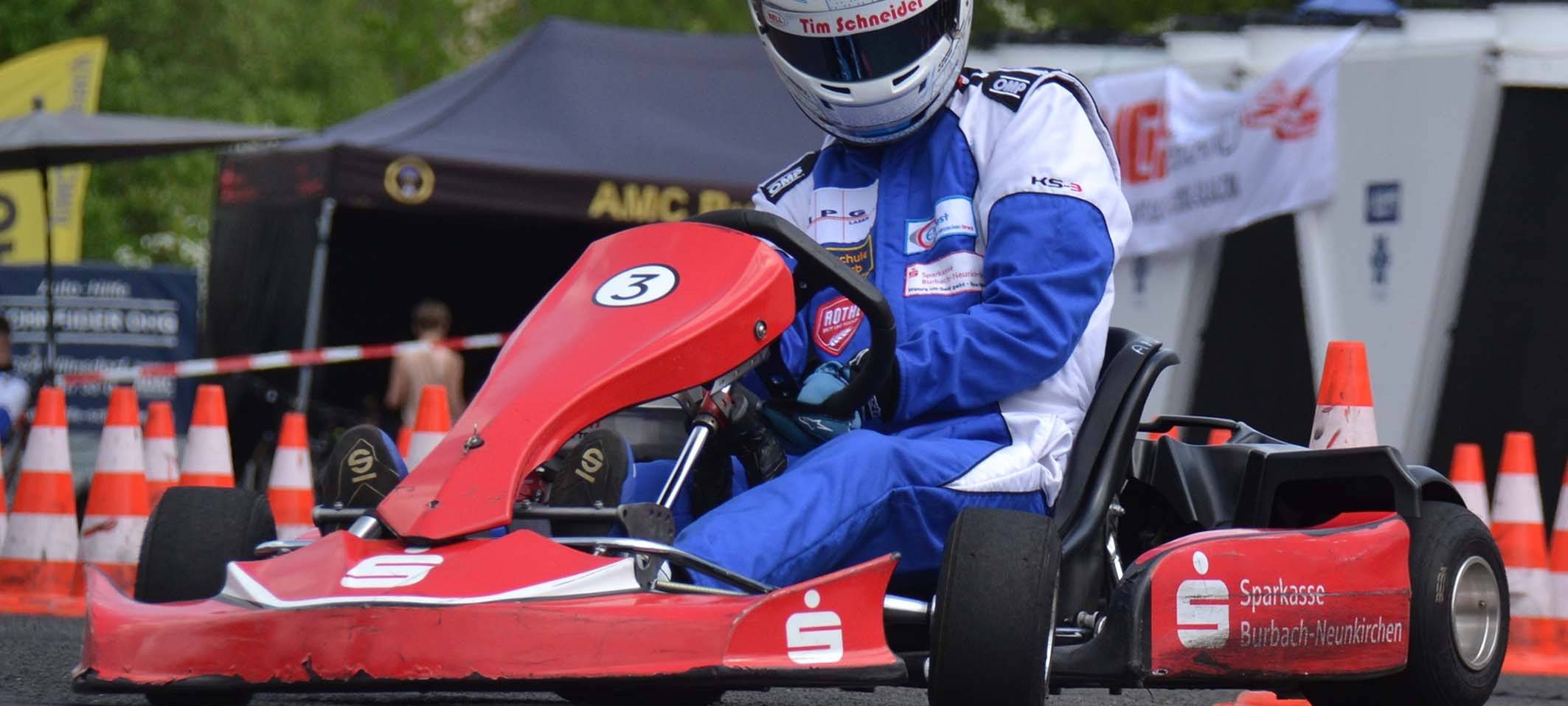 Ein rotes Kart auf einer geteerten Strecke. Ein Jugendlicher mit einem weißen Helm lenkt das Kart. Ein reifen ist leicht in der Luft.