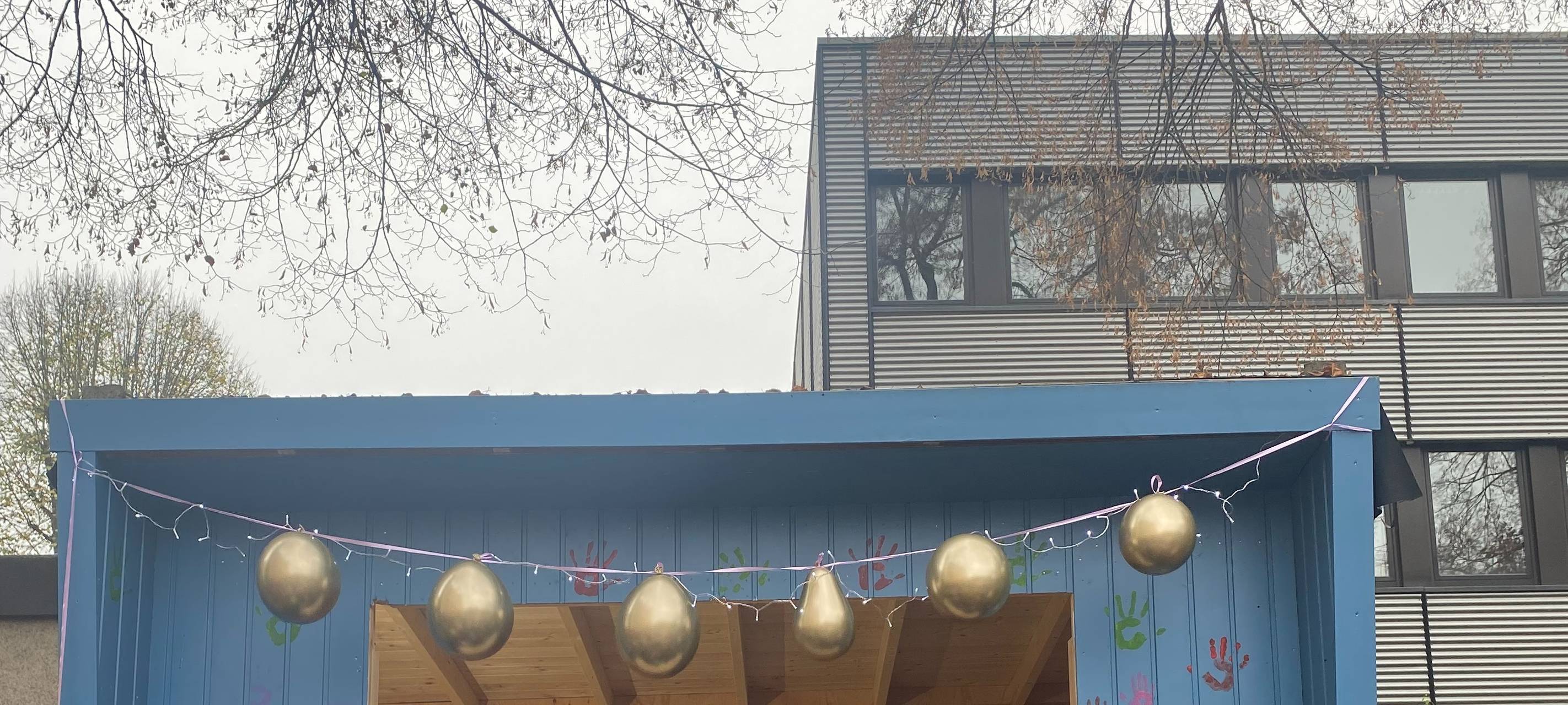 Viele Kinder vor einer blauen Hütte auf einem Schulhof. In der Hütte stehen Menschen und an der Wand hängen Springseile, Hula-Hoop-Reifen