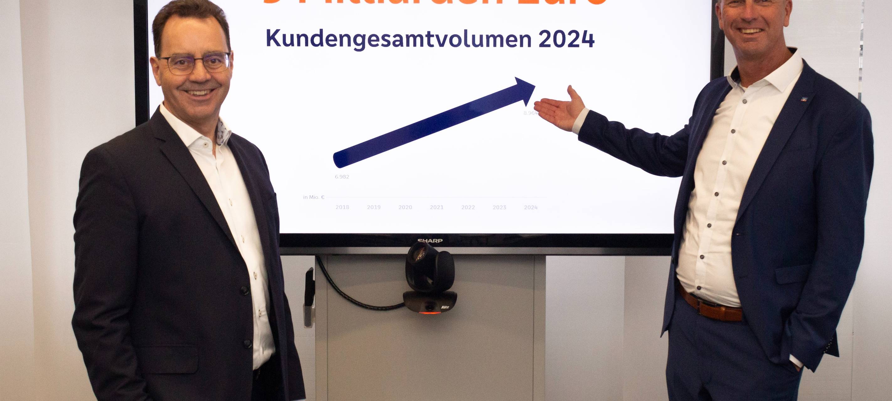 Zwei Männer von der Volksbank im Anzug stehen vor einem Fernseher.